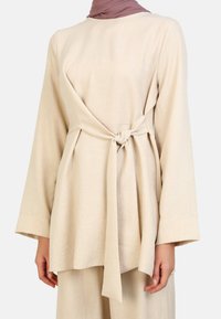 Beige linen top with long, wide sleeves, featuring a front knot detail at the waist and a straight cut. Smooth texture and simple design.