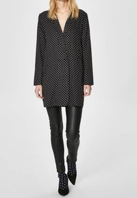 Schwarzes Polka-Dot-Hemdkleid mit V-Ausschnitt, Knopfverschluss vorne, kombiniert mit schwarzen Lederhosen und schwarzen Socken mit weißen Polka-Dots.