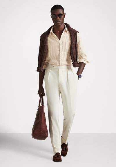 Άνδρας με γυαλιά ηλίου, μπεζ ριγέ πουκάμισο, κρεμ παντελόνι, καφέ πουλόβερ περασμένο πάνω από τους ώμους, κρατώντας καφέ δερμάτινη τσάντα, περπατώντας προς τα εμπρός.