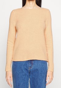 Pfirsichfarbener, gerippter Strickpullover mit langen Ärmeln und weitem Ausschnitt, kombiniert mit mittelhohen blauen Jeans. Weiche Textur.