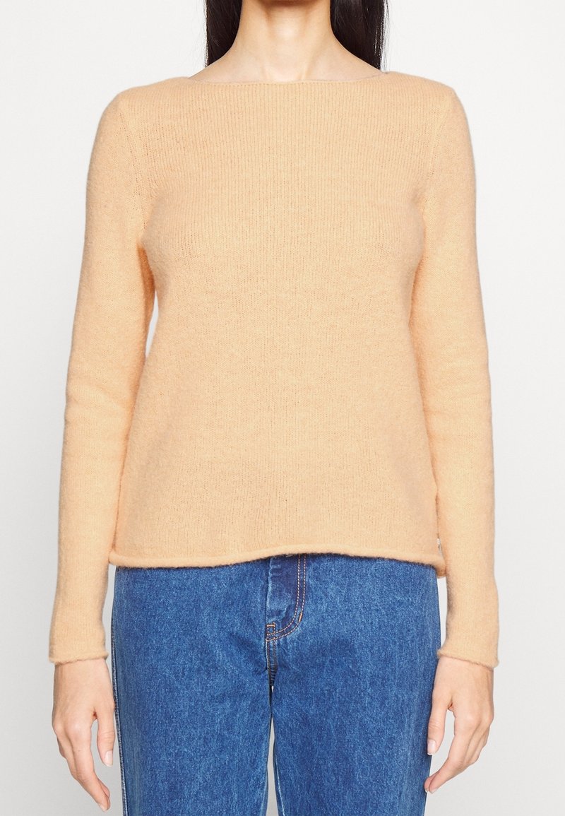 Pfirsichfarbener, gerippter Strickpullover mit langen Ärmeln und weitem Ausschnitt, kombiniert mit mittelhohen blauen Jeans. Weiche Textur.