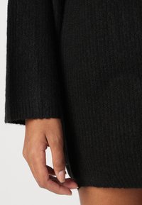 Black knit sweater with a ribbed texture, long sleeves, and a straight cut hem, with a hand positioned near the lower edge.