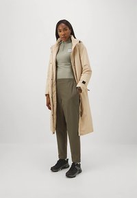 Casaco comprido bege por cima de uma blusa cinza e calças verdes-oliva. Ténis pretos com solas texturizadas. O casaco tem capuz e fechos de pressão.