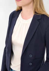 Granatowa marynarka wykonana z gładkiego materiału, z dwoma guzikami. Pod spodem lekka bluzka z dekoltem w falbanki.
