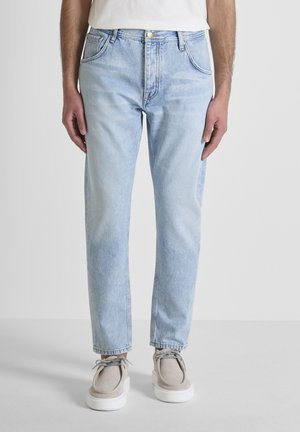 Hombre vestido con jeans de mezclilla azul claro, camisa blanca y zapatos beige con suelas gruesas blancas, posando sobre un fondo claro y liso.