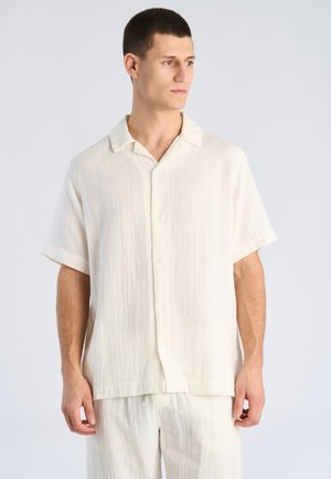 Młody mężczyzna w luźnej kremowej koszuli na krótki rękaw zapinanej na guziki i pasujących jasnych spodniach, patrzący w prawo.