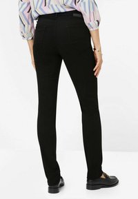 Schwarze dehnbare Jeans mit tailliertem Design, ausgestattet mit Gesäßtaschen und einer glatten Textur. Getragen mit einer gemusterten Bluse und Slippern.