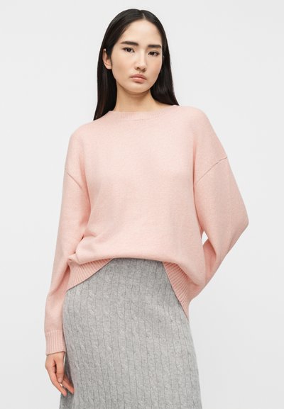 Γυναίκα με μακριά σκούρα μαλλιά, ντυμένη με ένα ροζ ανοιχτόχρωμο φαρδύ πουλόβερ και μια γκρι φουστάνι πλεχτό με μοτίβο καλώδιο, στέκεται μπροστά σε απλό φόντο.