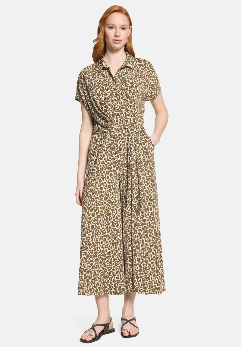 Femme aux cheveux roux portant une robe midi à manches courtes, ceinturée, beige et marron à imprimé léopard, et des sandales plates noires, se tenant devant un fond uni.