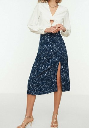 Witte top met lange mouwen en een keyhole-detail aan de voorkant, gecombineerd met een marineblauwe midirok met een wit stippenpatroon en een split aan de zijkant. Beige sandalen met bandjes.