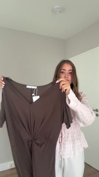 Hnedé šaty s krátkym rukávom, výstrihom do V a detailom twist v oblasti pása. Tkanina vyzerá mäkká a hladká, s jednoduchým, jednoliatym dizajnom.