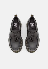 Mocassins en cuir noir avec pompons, présentant une surface texturée, des coutures jaunes et une semelle intérieure rembourrée. Marque Dr. Martens sur la semelle intérieure.