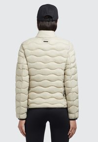 Jachetă puffer bej cu un design ondulat și quiltat, guler înalt și manșete elastice. Prezintă o mică etichetă cu logo pe spate. Purtați o șapcă neagră.