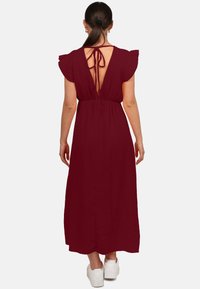 Abito maxi bordeaux in tessuto texturizzato, con maniche a sbuffo, schiena scoperta e chiusura con lacci. Indossato con sneakers bianche.
