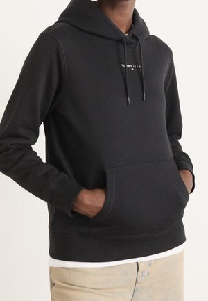 Osoba nosząca czarną bluzę z kapturem i kieszenią z przodu oraz logo "Tommy Jeans", ręce w kieszeniach, beżowe spodnie, biała koszula pod spodem.