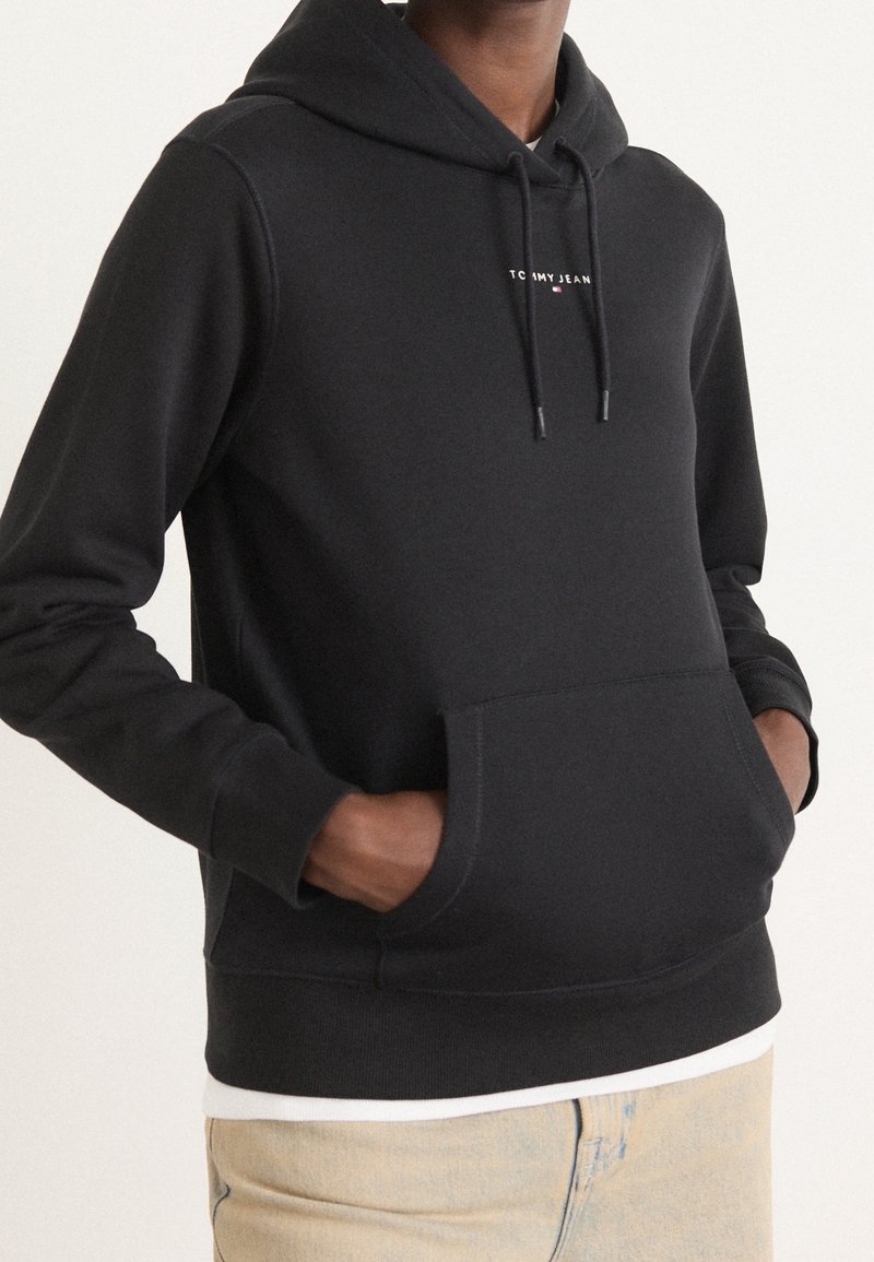 Personne portant un sweat à capuche noir avec poche avant et logo "Tommy Jeans", mains dans les poches, pantalon beige, chemise blanche en dessous.