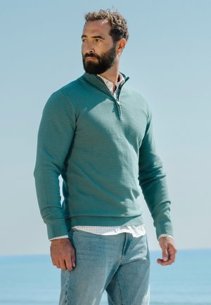 Bărbat cu barbă, purtând un pulover verde-teal cu fermoar până la piept peste o cămașă albă și blugi deschiși, stând lângă o mare liniștită sub un cer albastru senin.