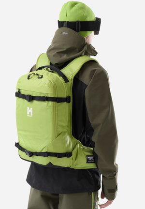 Personne portant un bonnet vert, une veste vert olive et un sac à dos Millet vert vif avec des sangles noires, vue de dos sur un fond blanc.