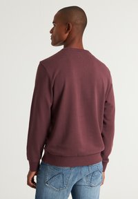 Sudadera burdeos con cuello redondo y puños acanalados. Presenta un acabado texturizado y un sutil detalle en la parte posterior. Combinada con jeans azules.