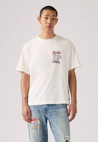 Νεαρός άνδρας που φοράει ένα λευκό μπλουζάκι με γραφικά από την ταινία Toy Story και φθαρμένα ανοιχτόχρωμα τζιν, έχοντας ως φόντο μία απλή επιφάνεια.