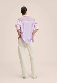 Hellviolette und rosa gestreifte Bluse mit hochgekrempelten Ärmeln, kombiniert mit cremefarbenen Hosen. Der Stoff wirkt leicht und die Passform locker.