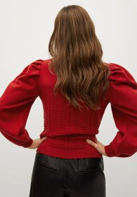 Pull en tricot rouge avec des manches bouffantes, motif texturé au dos, ourlet côtelé, porté avec un pantalon en cuir noir.