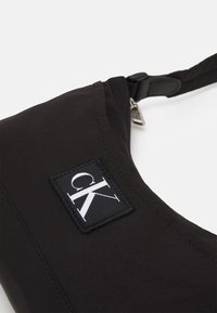 Musta kangas olkalaukku, jossa on teksturoitu pinta, kirjailut ja näkyvä CK-logo suorakaiteen muotoisessa kuviossa.