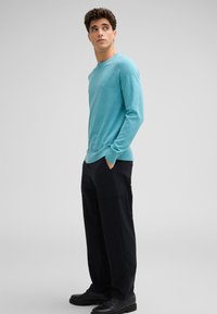 Türkiser Pullover mit langen Ärmeln, gerippten Bündchen und Saum, kombiniert mit lockeren schwarzen Cargo-Hosen und schwarzen Schuhen, Model steht seitlich.