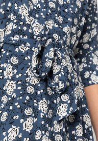 Blommig klänning i blått tyg med vita och svarta mönster. Har ett knytmidja och knappdetalj, med korta ärmar och en andningsbar textur.