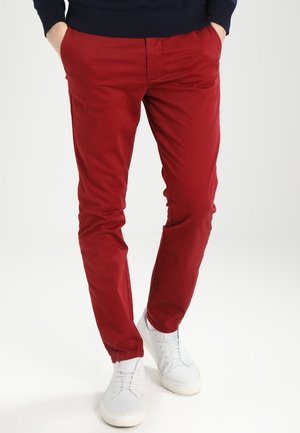 Mann trägt eng anliegende rote Hosen, weiße Sneakers und einen dunkelblauen Pullover, steht mit den Händen in den Taschen vor einem einfarbigen Hintergrund.