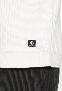 Weißer Strickpullover mit gerippter Textur und einem schwarzen Markenlabel mit geometrischem Logo. Kombiniert mit einem dunklen, gerippten Stoff darunter.