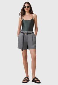 Bañador de una sola pieza en verde oscuro mate con tirantes ajustables con acentos dorados, combinado con pantalones cortos grises a medida que presentan un cinturón trenzado negro.