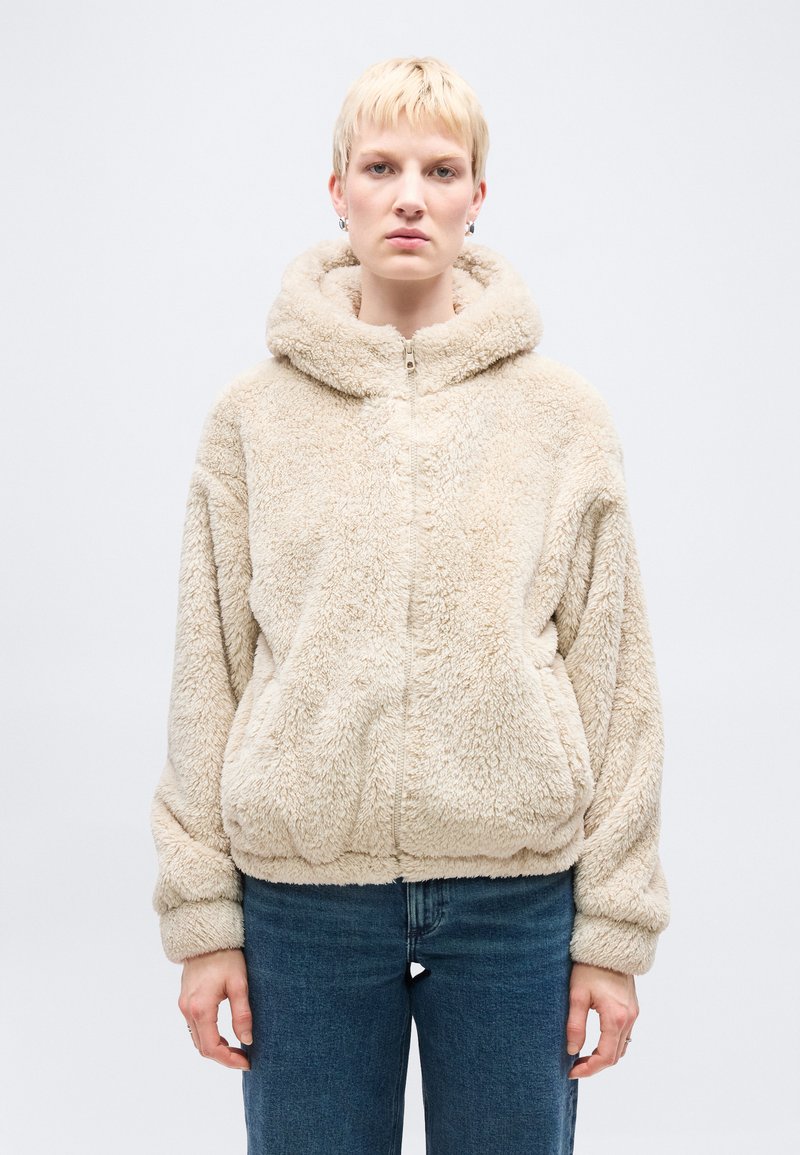 Άτομο με κοντά ξανθά μαλλιά, φοράει μπεζ χνουδωτό φούτερ με φερμουάρ και κουκούλα, και μπλε τζιν, στέκεται μπροστά σε λευκό φόντο.