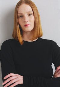 Maglione nero a maniche lunghe con collo rotondo, texture liscia e design aderente. Il modello presenta capelli lunghi e dritti, fino alle spalle, con le braccia incrociate.