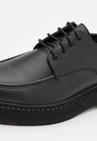 Scarpa in pelle nera con una finitura liscia, punta rotonda e sei occhielli per i lacci. La suola presenta una gomma testurizzata per una buona presa.