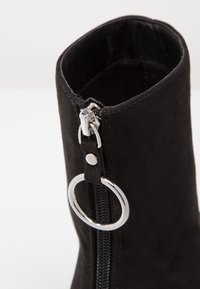 Bottine en daim noire avec une fermeture éclair en métal argenté et un accent en métal rond, au toucher lisse et au design ajust�é.