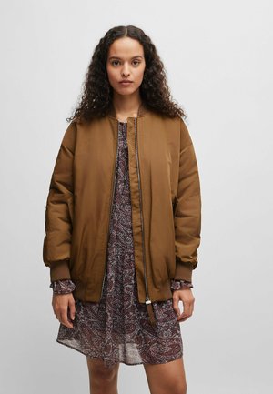 Braune Bomberjacke mit gerippten Bündchen und Reißverschluss, getragen über einem floral gemusterten Kleid mit Rüschenschalen.