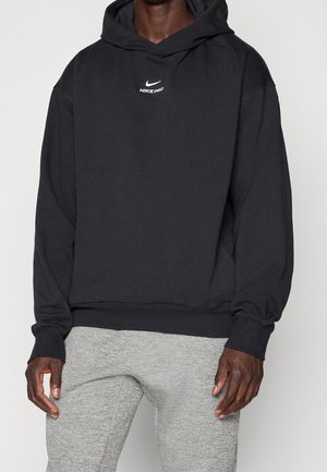 Personne portant un sweat à capuche Nike Pro noir avec un logo blanc sur la poitrine et un pantalon de survêtement gris clair, debout devant un fond uni.