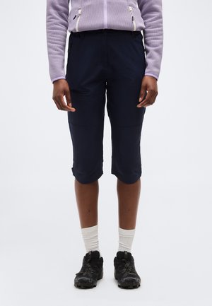 Άτομο που φοράει μαύρο σορτς μέχρι το γόνατο με τσέπες με φερμουάρ, λευκές κάλτσες μέχρι τον αστράγαλο, μαύρα αθλητικά παπούτσια και ανοιχτόμωβ ζακέτα με φερμουάρ.