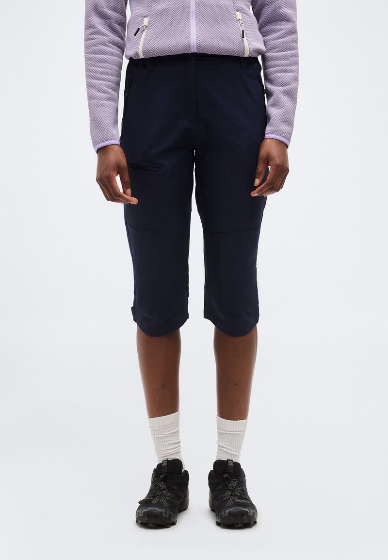 Άτομο που φοράει μαύρο σορτς μέχρι το γόνατο με τσέπες με φερμουάρ, λευκές κάλτσες μέχρι τον αστράγαλο, μαύρα αθλητικά παπούτσια και ανοιχτόμωβ ζακέτα με φερμουάρ.