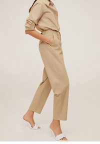 Un pantalon beige taille haute, coupe droite, avec des poches latérales, associé à une chemise boutonnière marron clair et des sandales à talons blancs.