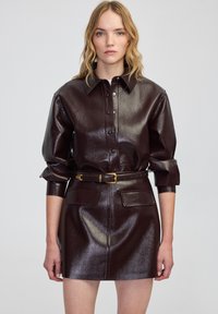 Vestido camisero de cuero burdeos con botones en la parte delantera, mangas largas y cintura ceñida con cinturón. Presenta dos bolsillos frontales, textura suave y acabado brillante.