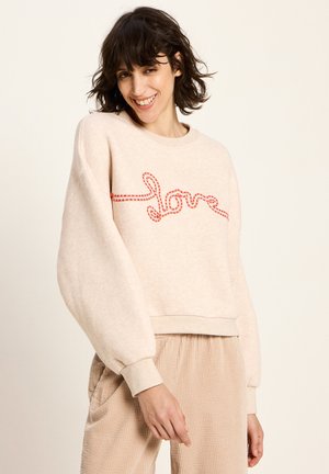 Frau mit kurzem dunklem Haar, die lächelt und einen beigen Pullover mit rotem "Liebe"-Stich sowie passende beige Cordhosen trägt.