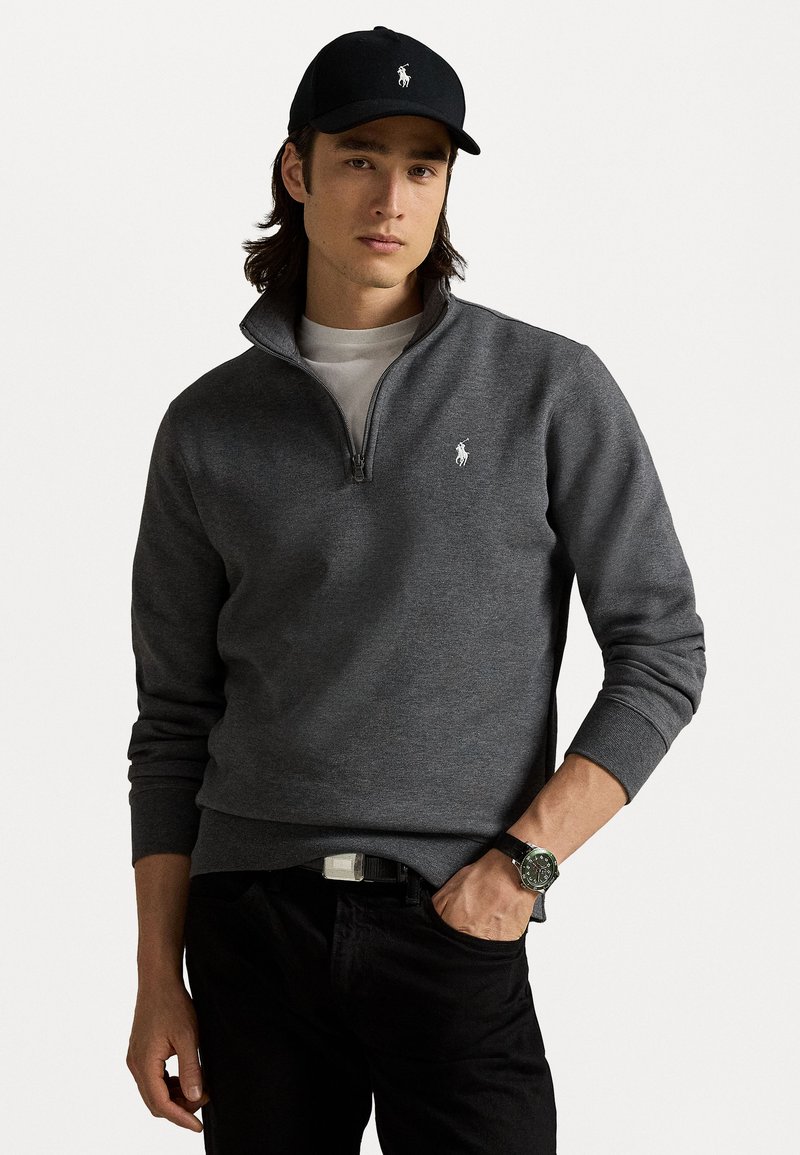 Grijze half-zip pullover trui met geribbelde manchetten, voorzien van een klein wit logo op de linkerborst, gedragen over een wit t-shirt en zwarte broek.