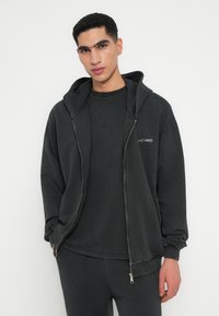Sudadera negra con cremallera y camiseta de cuello redondo negra a juego. Presenta una textura suave, un ajuste relajado y un detalle de logo minimalista.