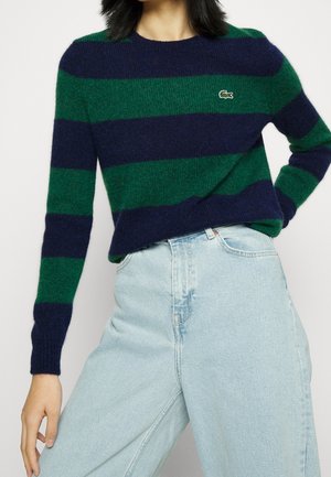 Femme portant un pull à larges rayures bleu marine et vert avec le logo Lacoste, associé à un jean taille haute bleu clair.