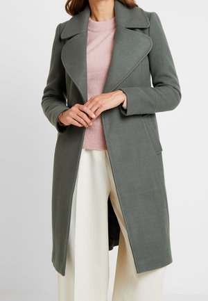 Femme portant un manteau long vert discret sur un pull rose et un pantalon large crème, debout les mains doucement jointes à la taille.