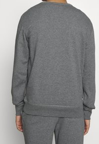 Muž v jednoduché šedé mikině s dlouhým rukávem a odpovídajících šedých kalhotách, zobrazený zezadu na bílém pozadí.