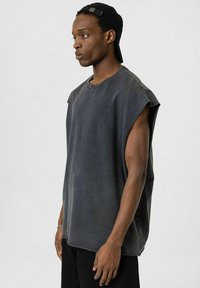 Mouwloze grijze stoffen shirt met een relaxed fit, ronde halslijn en ruwe randen, aangevuld met een persoon die een zwarte pet draagt.