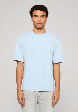 Mężczyzna o kręconych włosach, ubrany w jasnoniebieski t-shirt z krótkim rękawem oraz jasnoszare jeansy, stojący na jednolitym tle.